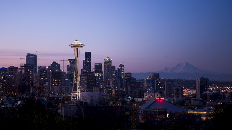 The image shows the Seattle skyline at twilight, featuring the Space Needle, numerous skyscrapers, and Mount Rainier in the background.