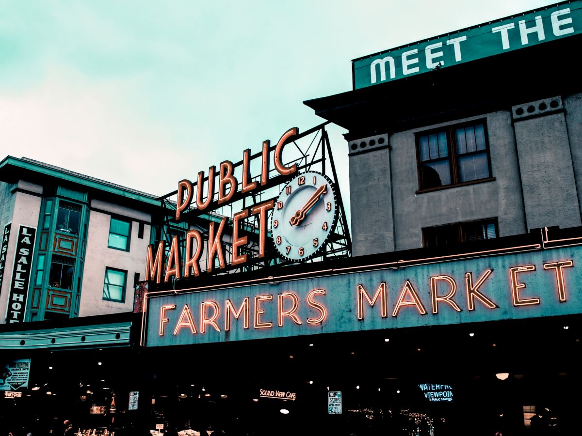 The image shows a Public Market with a neon "Farmers Market" sign and a clock under it, set against buildings.