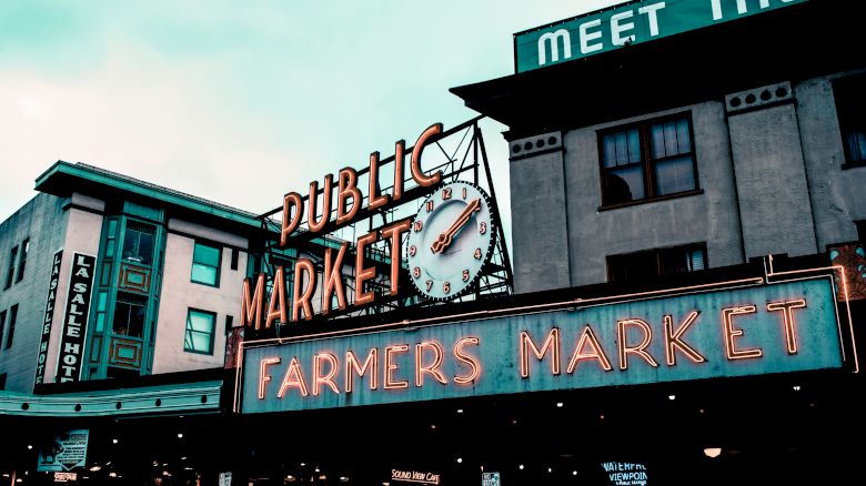 The image shows a Public Market with a neon "Farmers Market" sign and a clock under it, set against buildings.