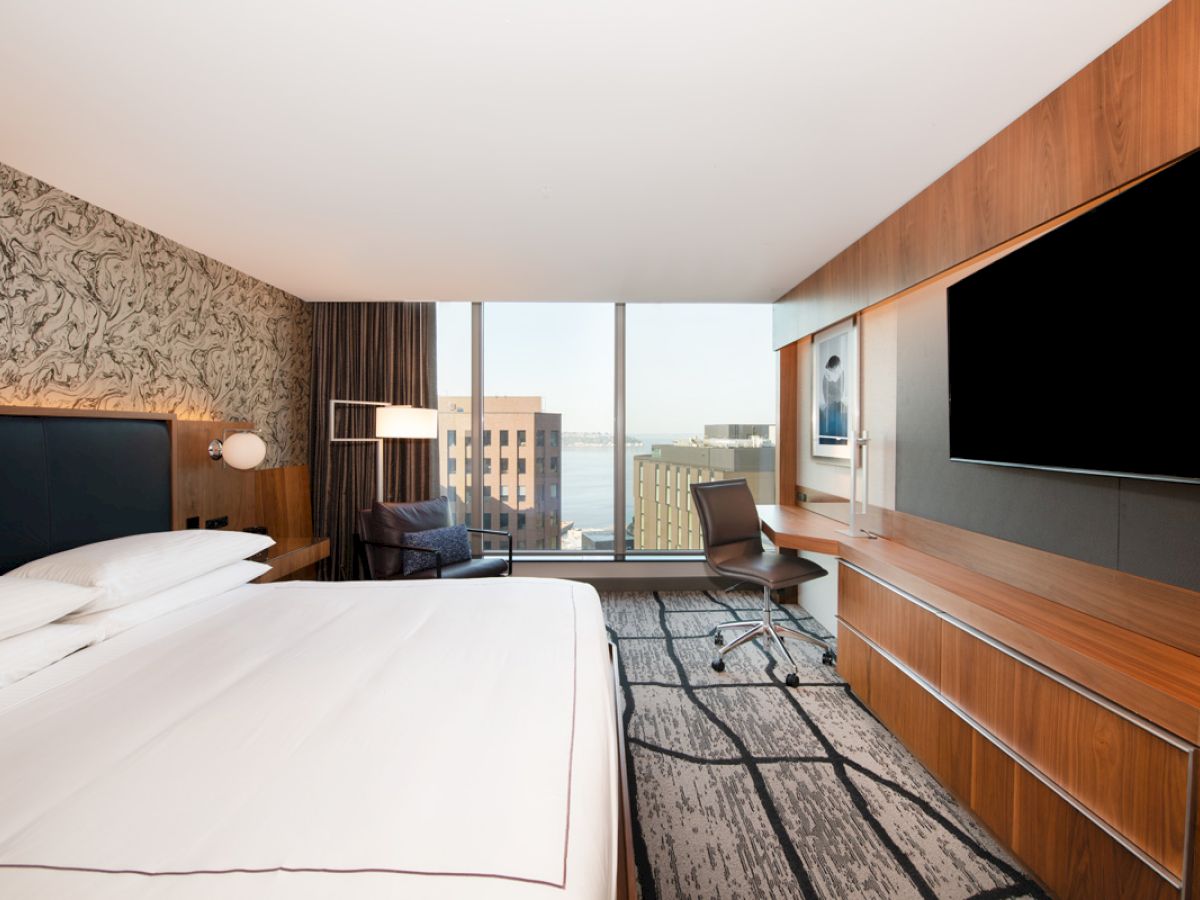 A modern hotel room with a large bed, a desk, a chair, a wall-mounted TV, and windows showcasing a cityscape view and partial body of water outside.