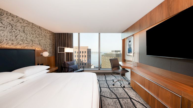 A modern hotel room with a large bed, a desk, a chair, a wall-mounted TV, and windows showcasing a cityscape view and partial body of water outside.