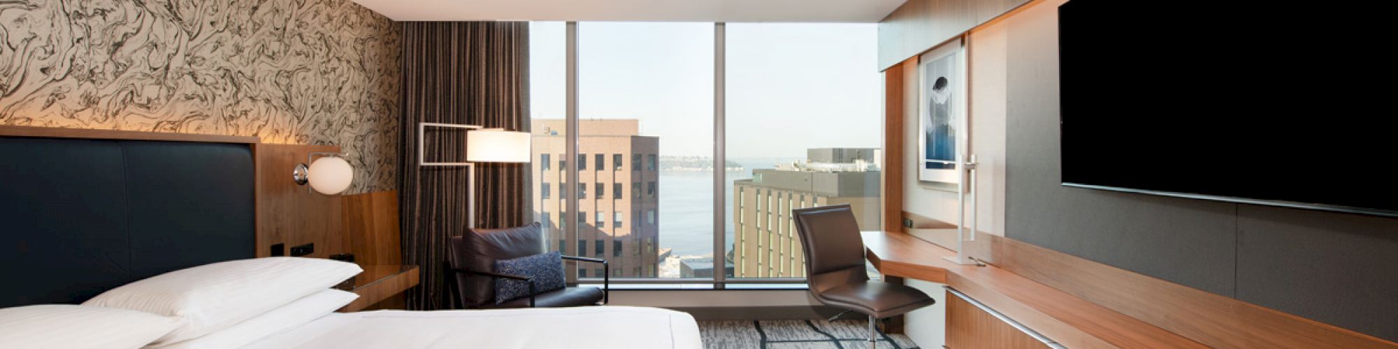 A modern hotel room features a large bed, wall-mounted TV, desk, chair, and window view of buildings and water in the distance.