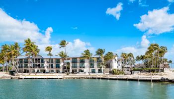 A beachfront building with multiple stories, palm trees, and clear blue skies by a calm body of water under a few scattered clouds.