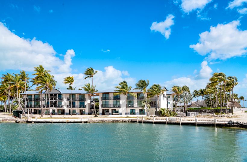 A beachfront building with multiple stories, palm trees, and clear blue skies by a calm body of water under a few scattered clouds.