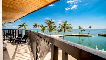 A balcony overlooking a serene beach with palm trees, clear blue water, and a bright sky with a few clouds.