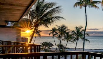 A sunset over a tropical beach with palm trees viewed from a wooden balcony, creating a serene and picturesque scene.