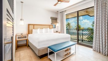 A bright and modern bedroom with a large bed, a ceiling fan, a view of palm trees, and a balcony overlooking a scenic beach setting.