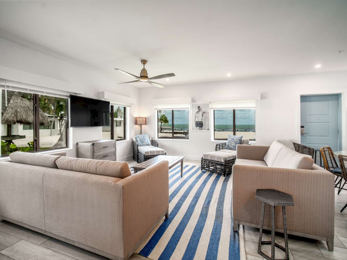 A modern living room with neutral sofas, a striped rug, and large windows revealing an ocean view. It has cozy seating and a ceiling fan.