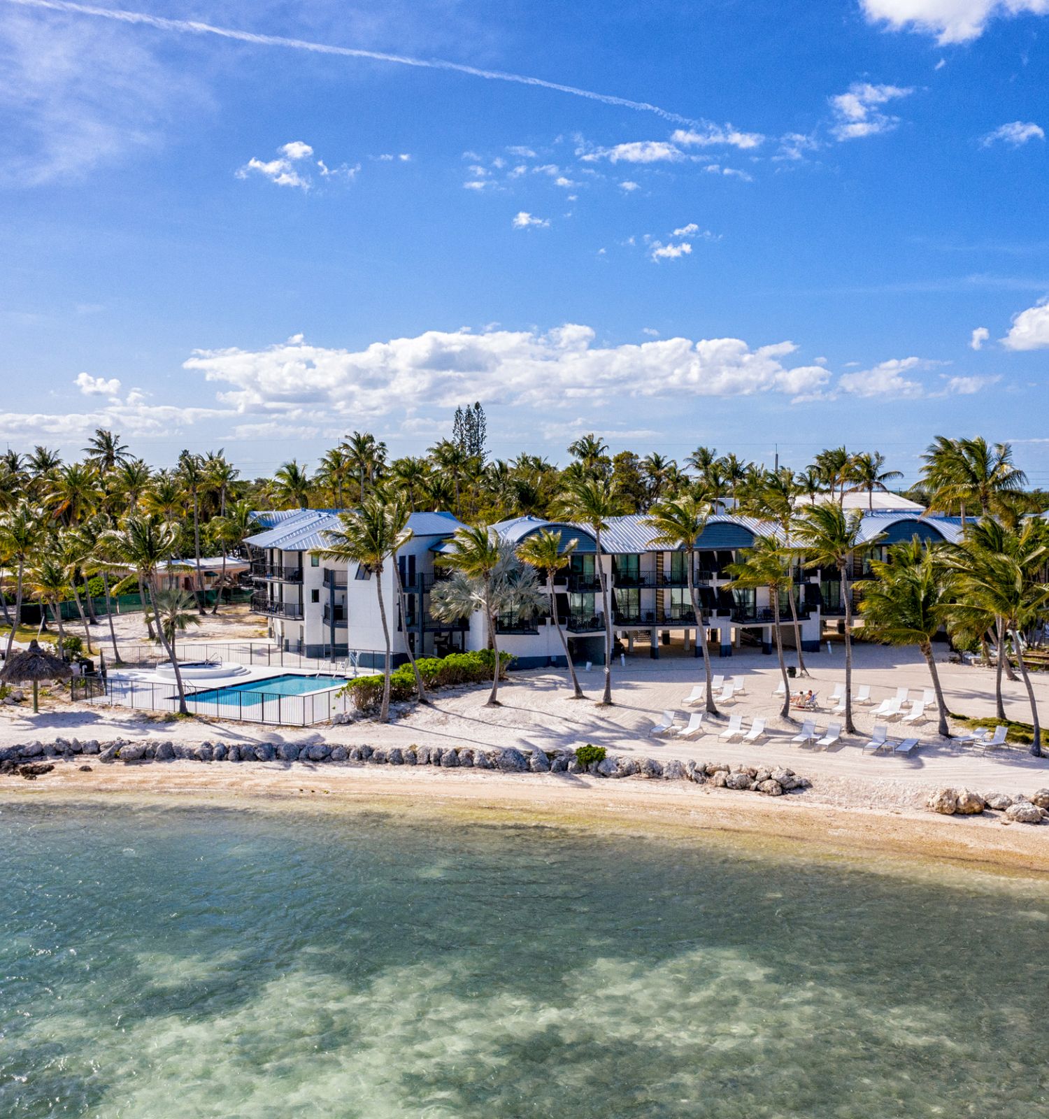 A picturesque beachfront property with palm trees, modern buildings, a pool, and a calm body of water in the foreground, on a sunny day.