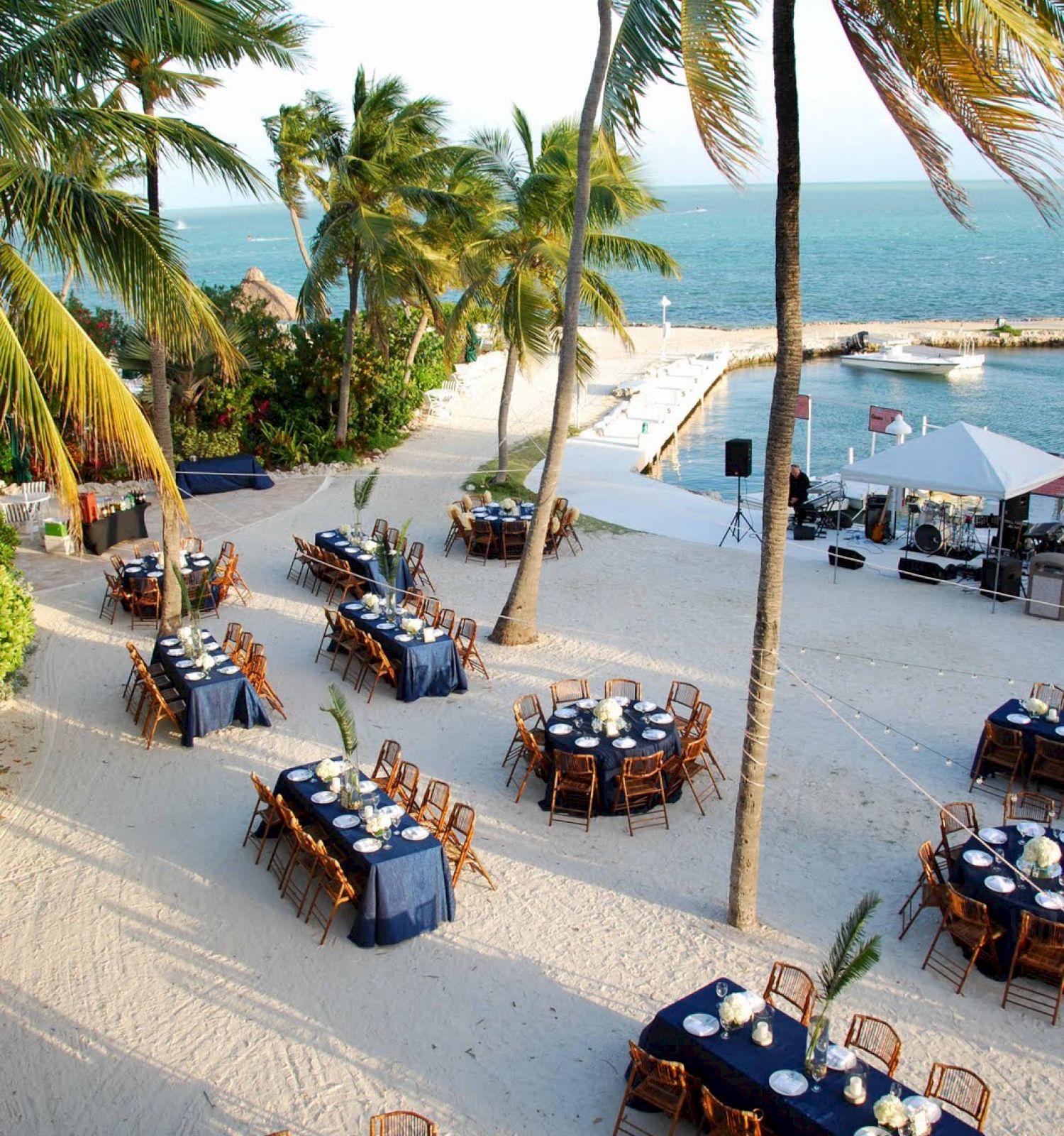 This image shows an outdoor event setup on a beach with round tables and chairs under palm trees, overlooking the ocean.