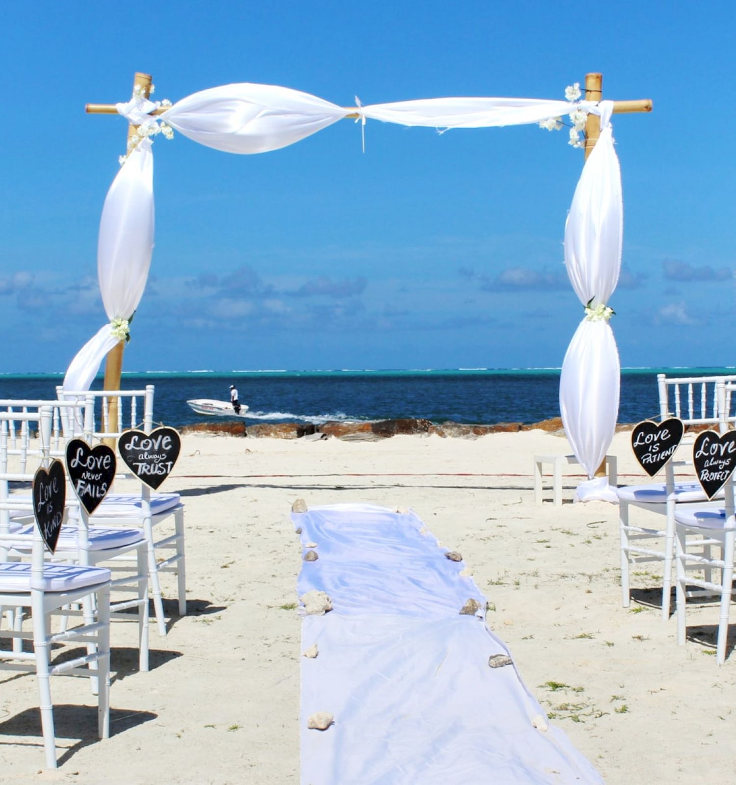 An outdoor beach wedding setup with white chairs, an aisle runner, a decorated arch, chalkboard signs, and palm trees against a sea backdrop.